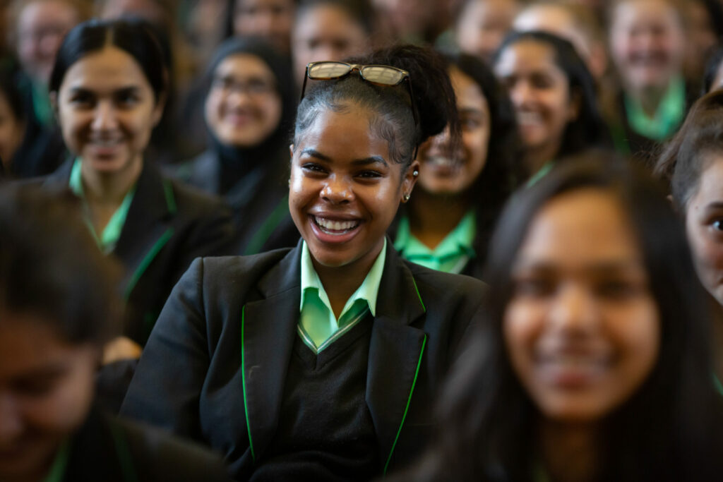 A female student smiling in a green and black uniform surrounded by her peers in assembly