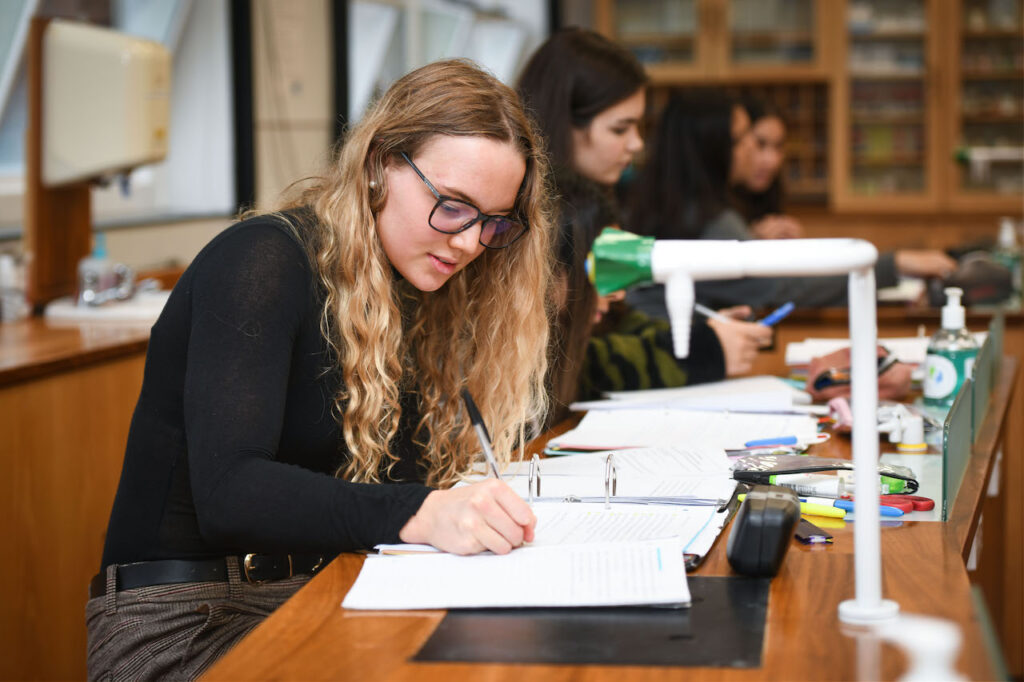 A female student writing notes at a desk in a classroom sat with her other peers
