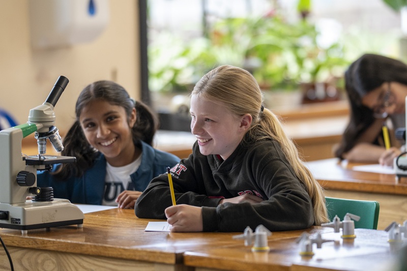 Two Thirds (Year 7s_ sitting at a wooden bench in a science lab, one of them holding a pencil and writing on a piece of paper as the other one smiles at the camera.
