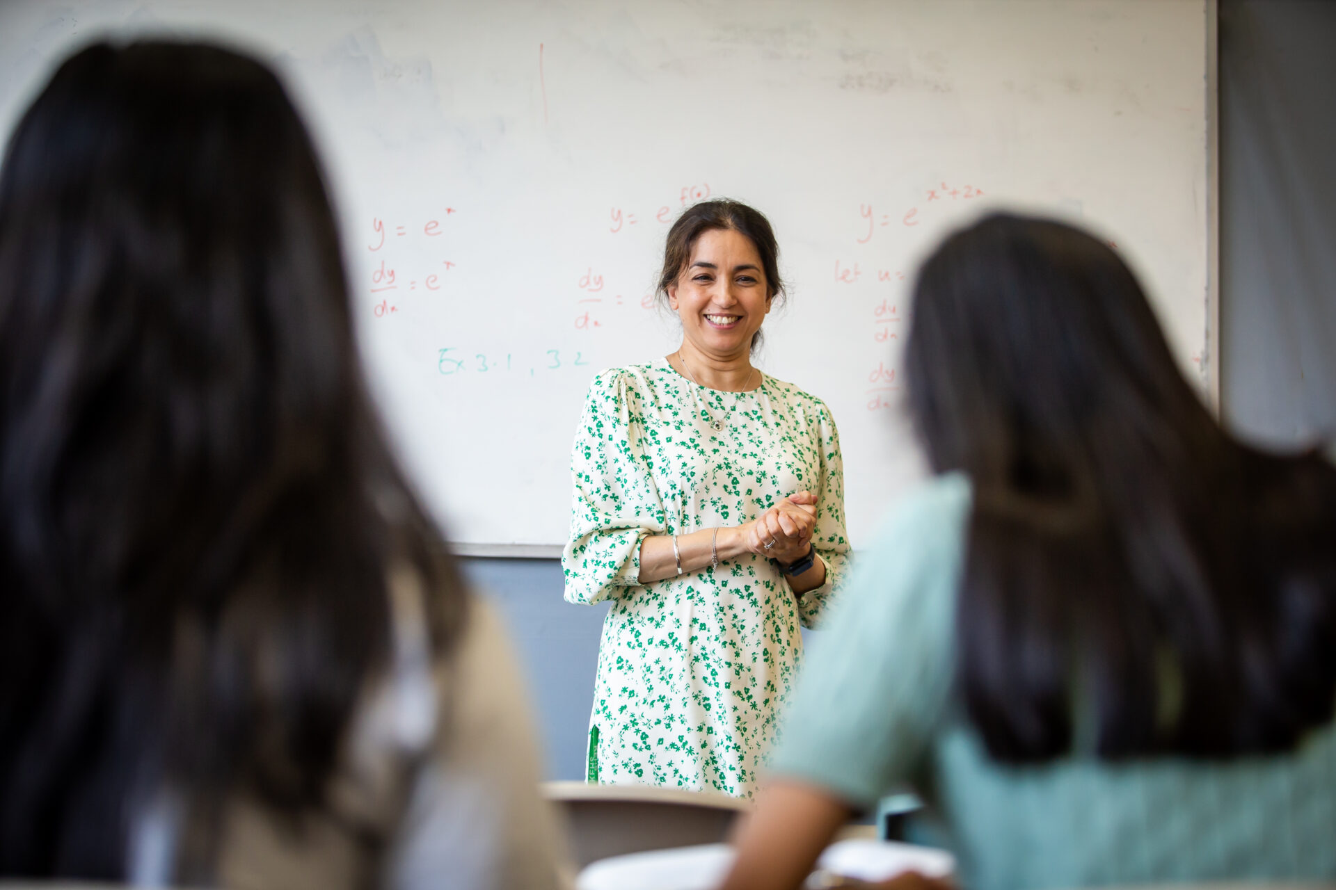 A teacher stands in front of a whiteboard with mathematical equations written on it, facing two students.