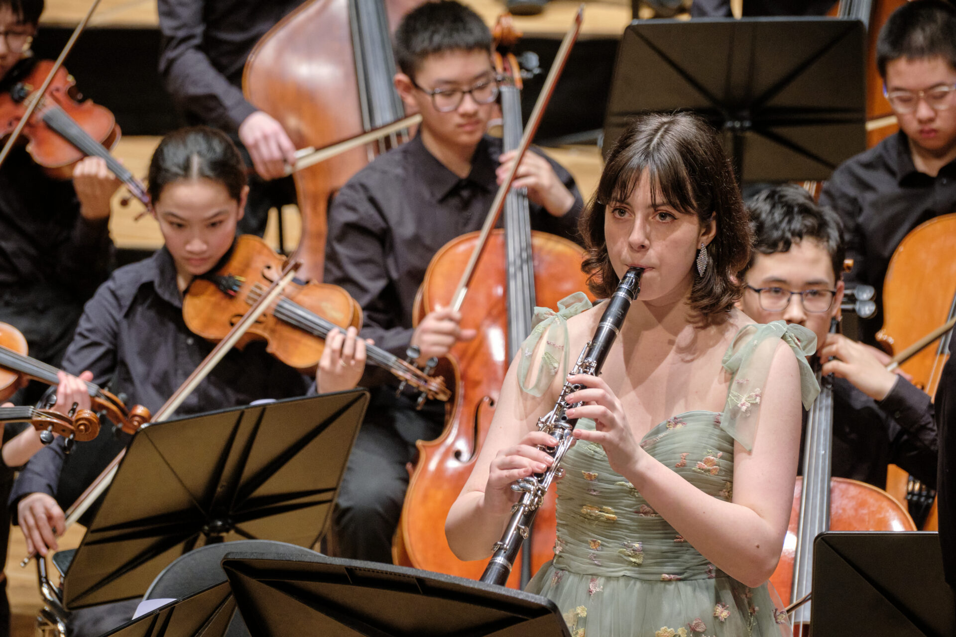A young girl plays the clarinet in an orchestra with other musicians playing violins and cellos behind her.