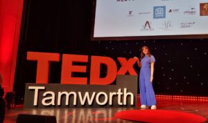 A pupil in a blue jumpsuit stands on a stage next to large TEDx Tamworth sign.