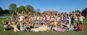 A large group of people covered in colourful powder are posing for a photo on a grassy field with their arms raised in celebration.