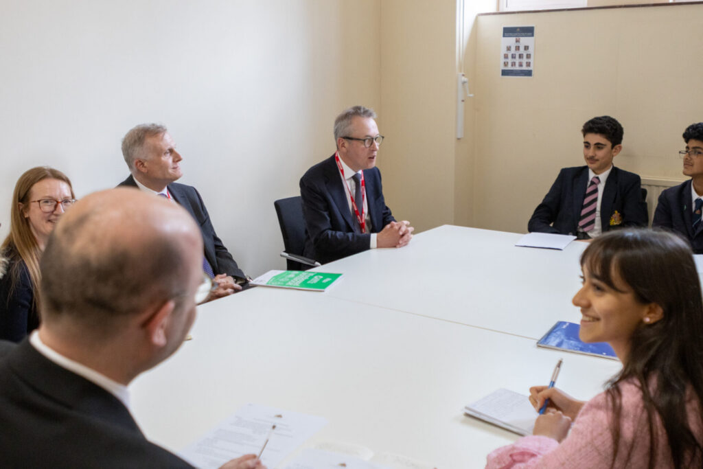 A group of adults and pupils are seated around a large white table in a conference room, engaged in a discussion. One adult is speaking while gesturing with his hands. pupils are taking notes.