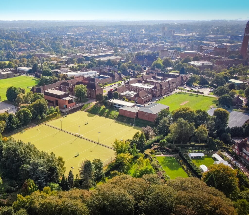 Aerial view of KEHS, with the green hockey astros in the foreground, the 1930s brick building in the centre, and the green sports field to the right. In the background are trees showy leafy south Birmingham and the university campus.