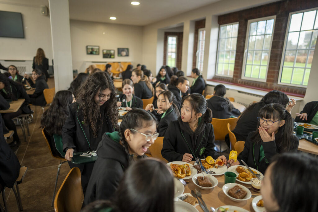 Students seated around a long dining table, eating and chatting in a bright school cafeteria.