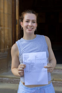 A student standing outdoors smiling and holding her exam results. 