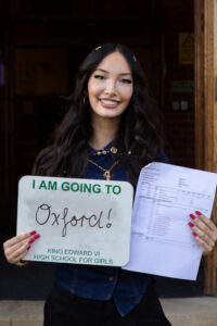 A smiling student holding her exam results and a sign saying “I AM GOING TO Oxford” in front of a building entrance.