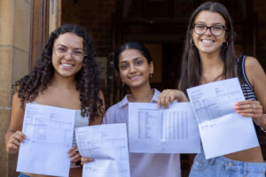 Three students outdoors smiling while holding printed exam result sheets and documents, standing in front of a brick building entrance.
