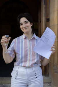 A student standing by a doorway holding her exam results in one hand and raising a small glass in the other. 