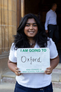 A student in a smiling while holding a sign that reads “I AM GOING TO Oxford,” standing outside a building. 
