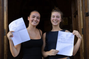 Two students smiling while holding up their examination results outside the school building. 