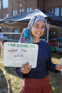 Smiling girl holding a sign that reads “I AM GOING TO explore the world outdoors near a building. 