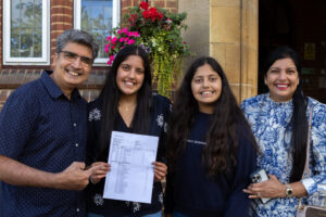 Four people smiling outdoors in front of a school building; one holds exam results.