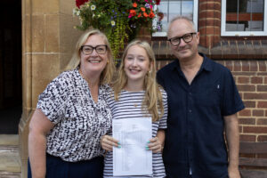 Three people stand smiling on steps in front of a brick building, one of the people is holding examination results.