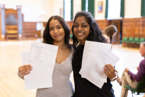 Two students smiling in a school hallway, holding up their exam results.