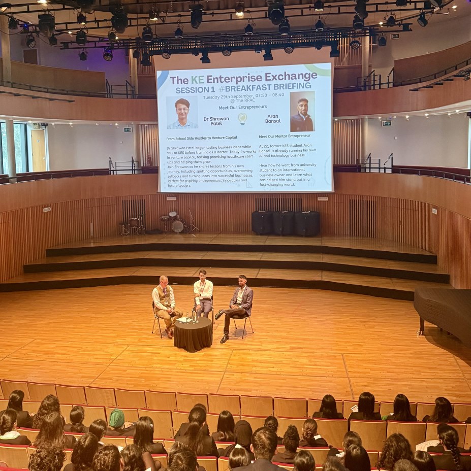 three speakers seated in front of an audience and a large screen behind them displaying “The KE Enterprise Exchange” and “Breakfast Briefing”.